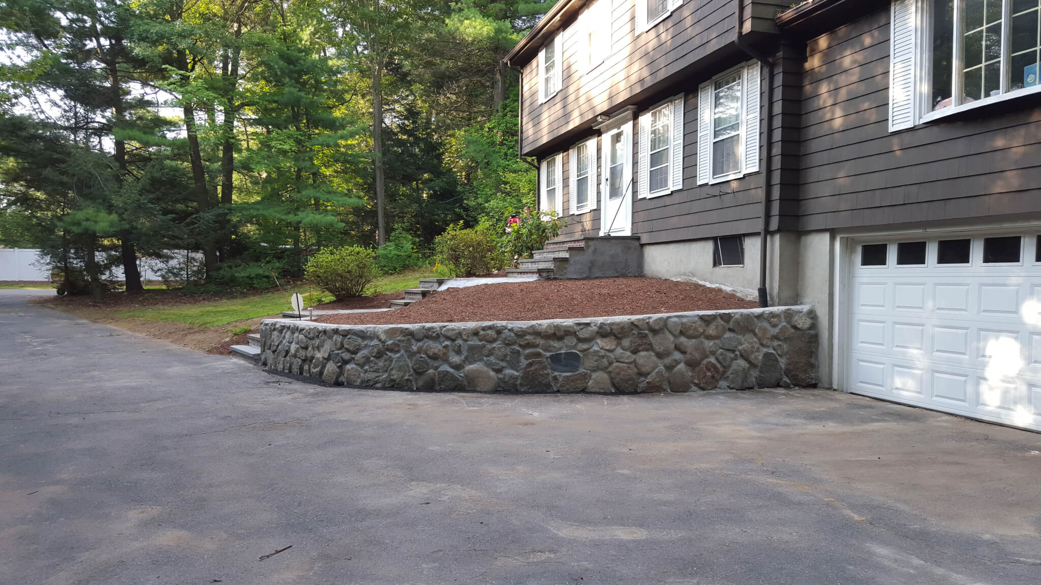 Stone retaining wall in front of a house, driveway, and landscaping. Dark siding and white garage door.