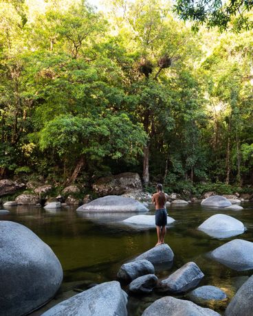 A person stands on a large boulder at Mossman Gorge, surrounded by mossy rocks and lush green trees.