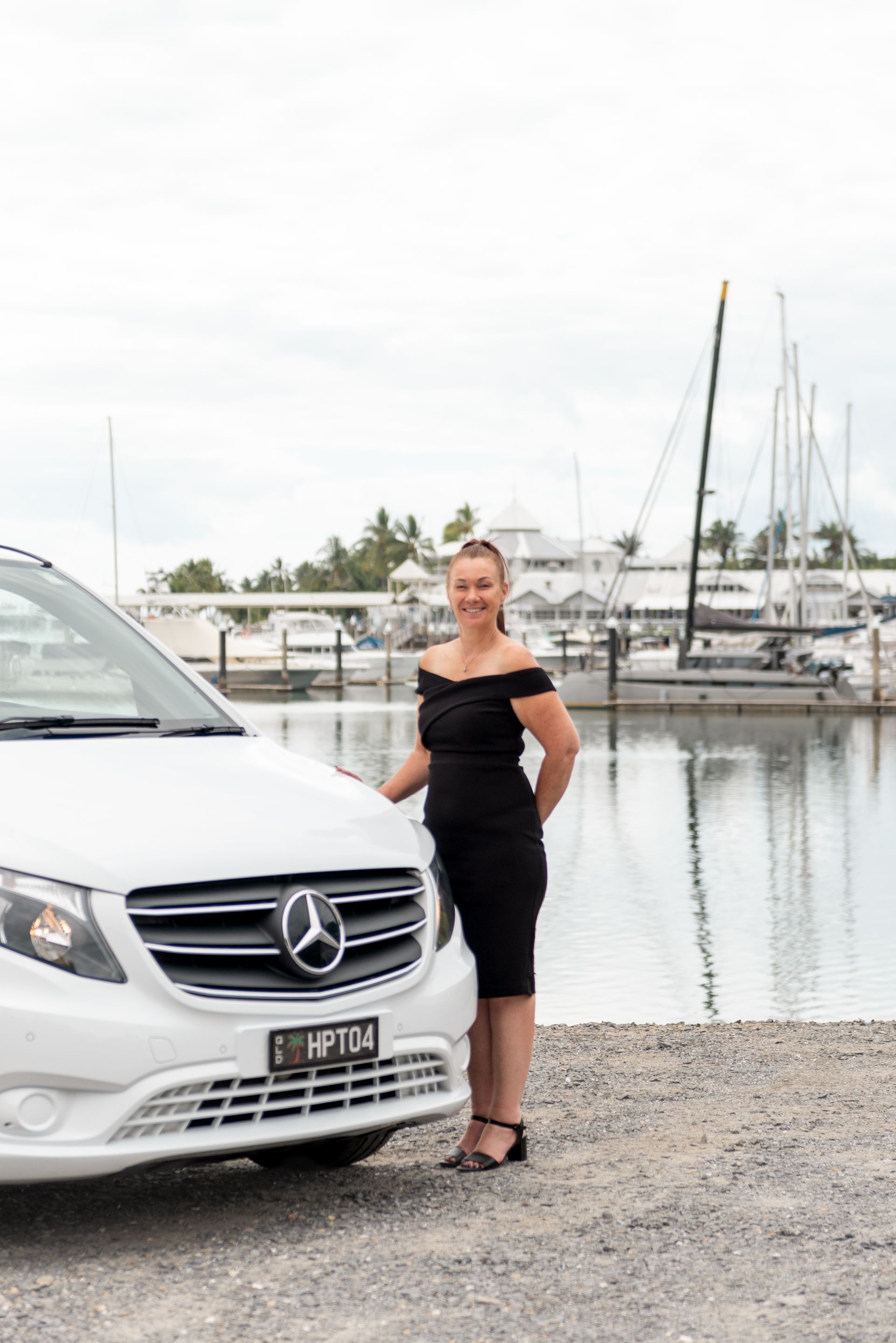 Heather in a black outfit stands beside a white Mercedes van parked at a boat marina in Port Douglas