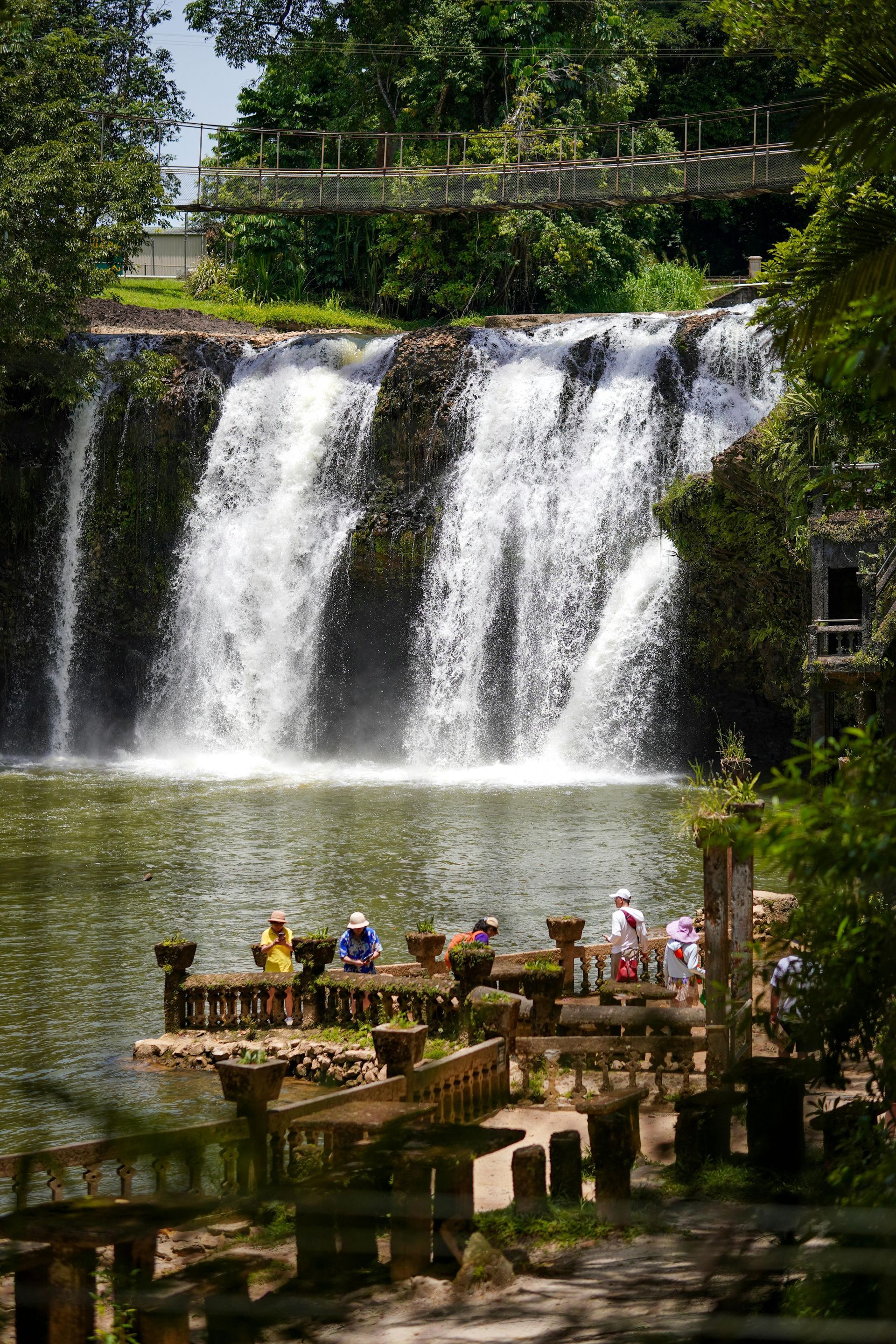 A wide, tiered waterfall cascades into a calm pool, with visitors on a terrace and a bridge above at Paronella Park