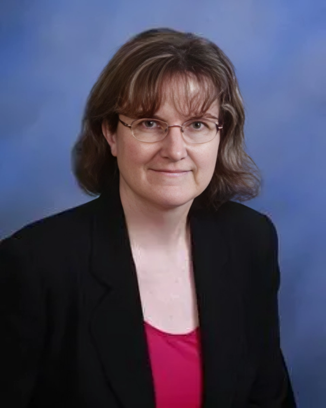 Woman with glasses, short brown hair, black blazer, and pink top. Blue background.