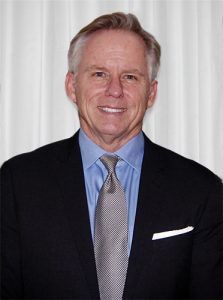 Man in blue shirt, grey tie, and black suit, smiling against a white backdrop.