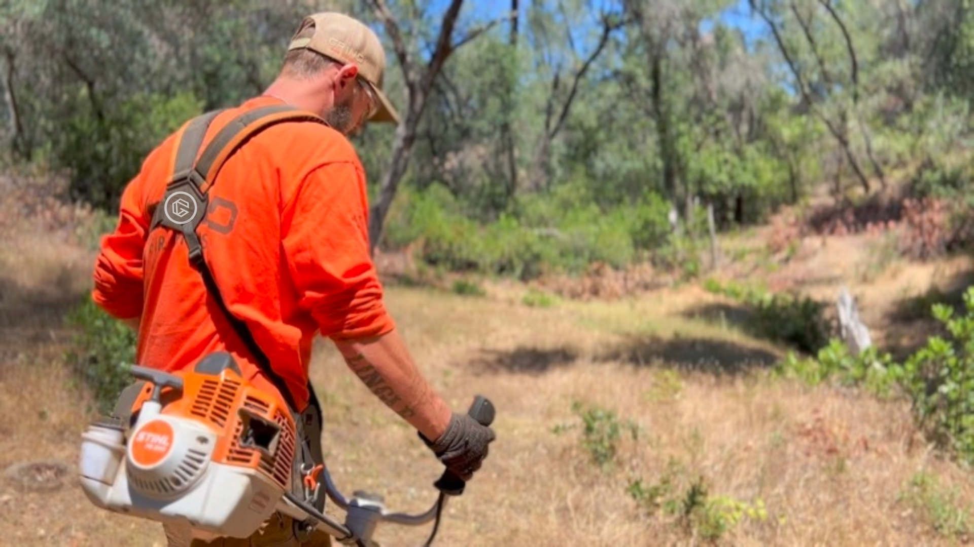 A person in Grind Fire Defense shirt using a weed whacker in a grassy, wooded area.