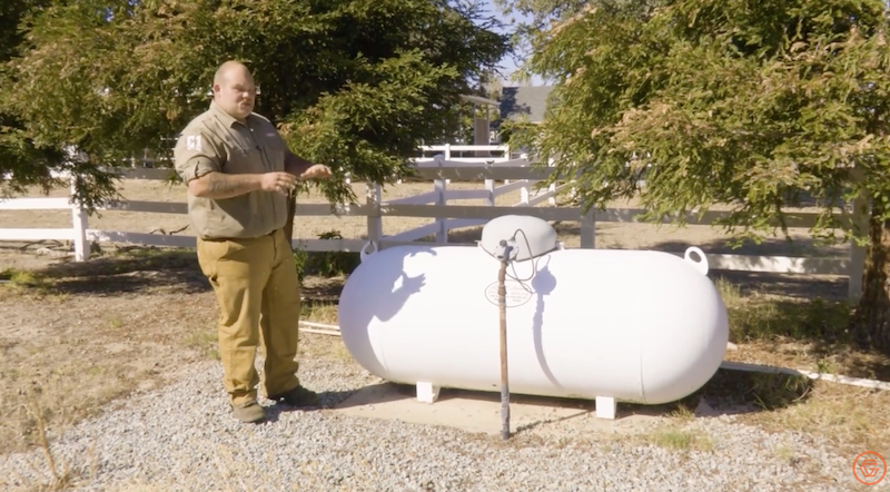 Man in uniform stands near a white propane tank outdoors, with a white fence and greenery in the background.
