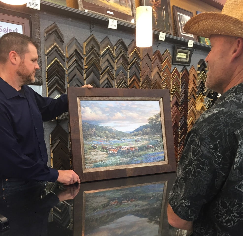 Two men examining a framed landscape painting in a frame shop. Various frame samples are in the background.
