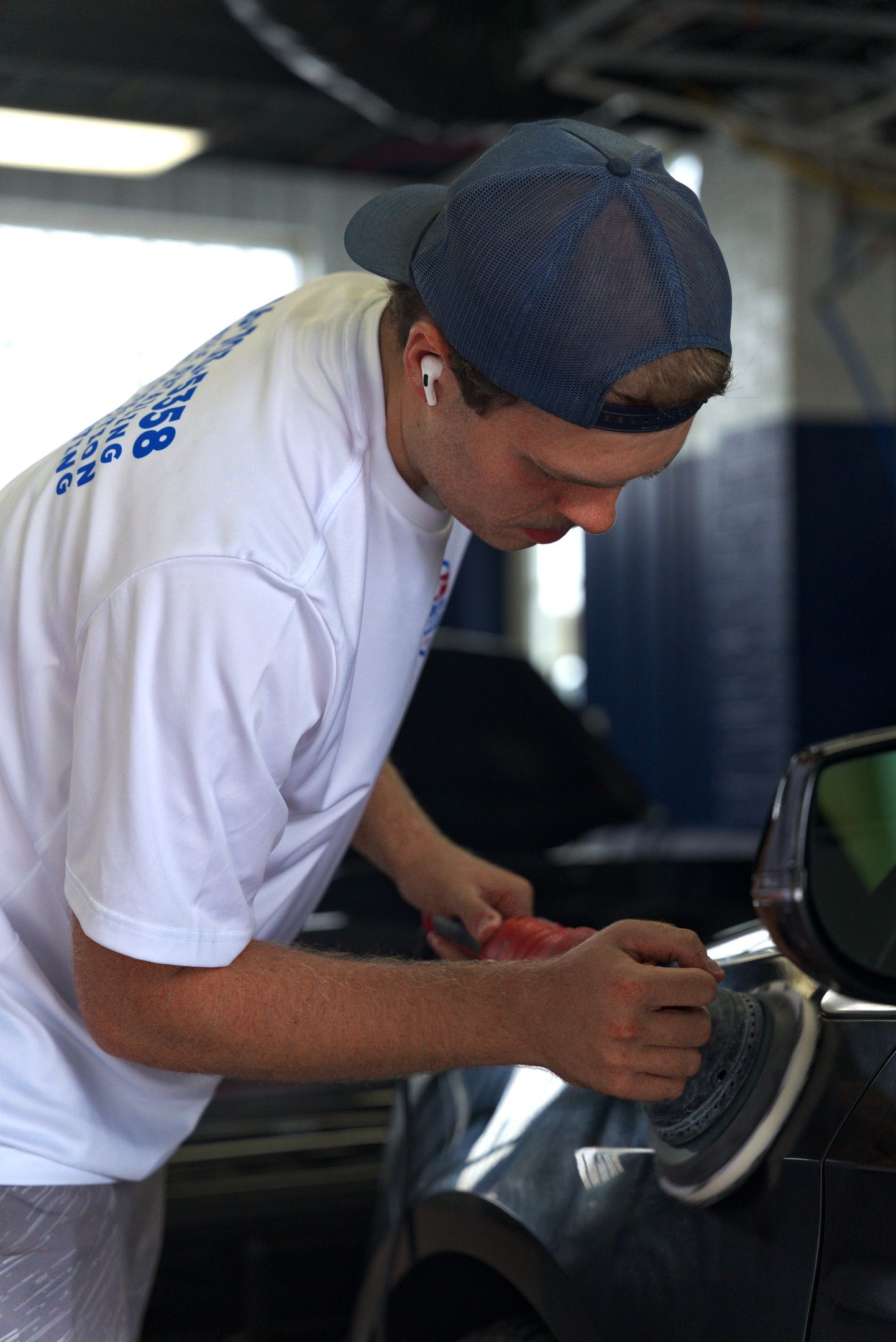 Worker in a white shirt and cap cleaning a car wheel in a garage.