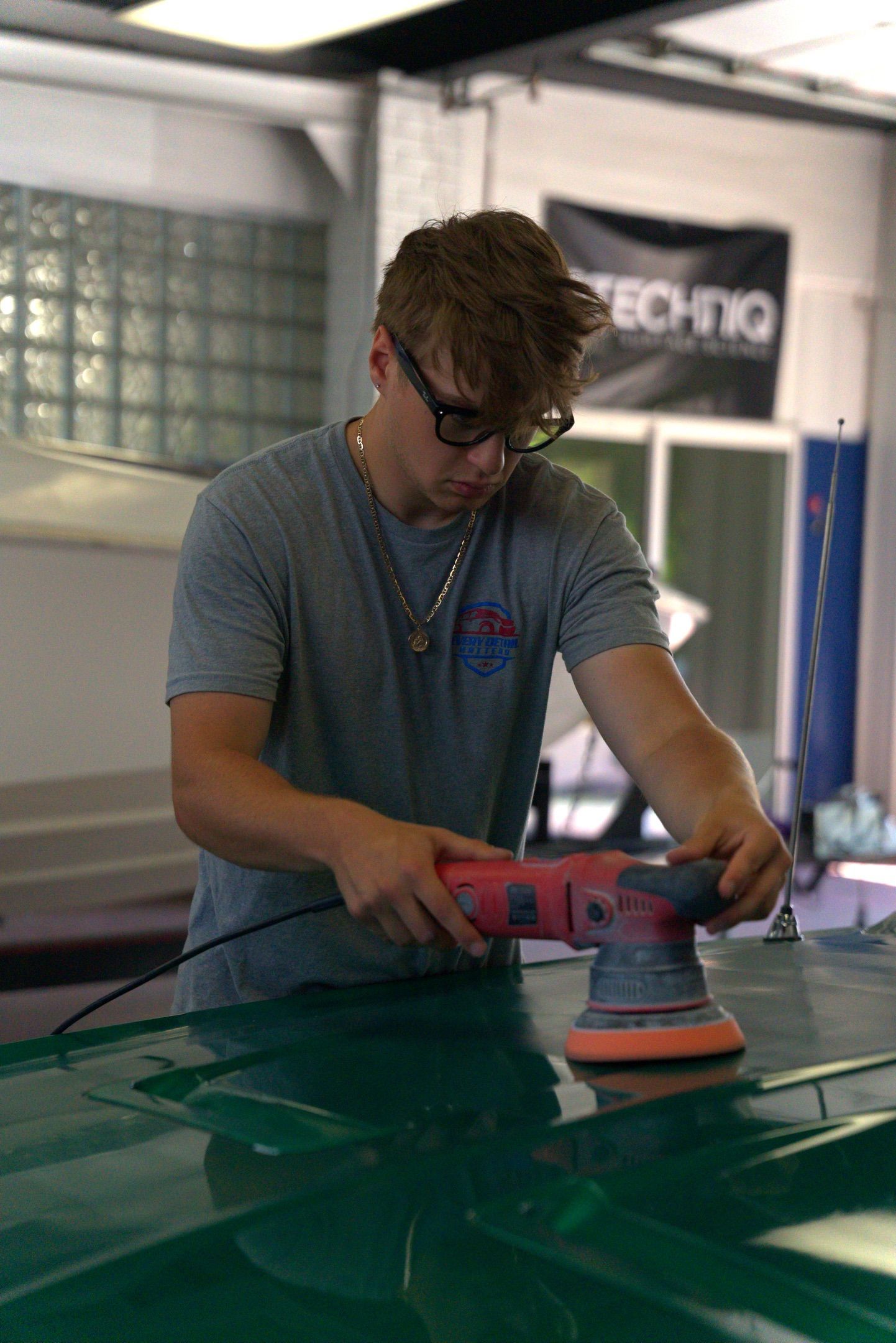 Person sanding a green car panel with an electric sander in a workshop.