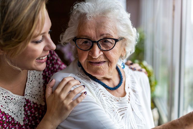 Young woman places arm on elder's shoulder, they look out a window.