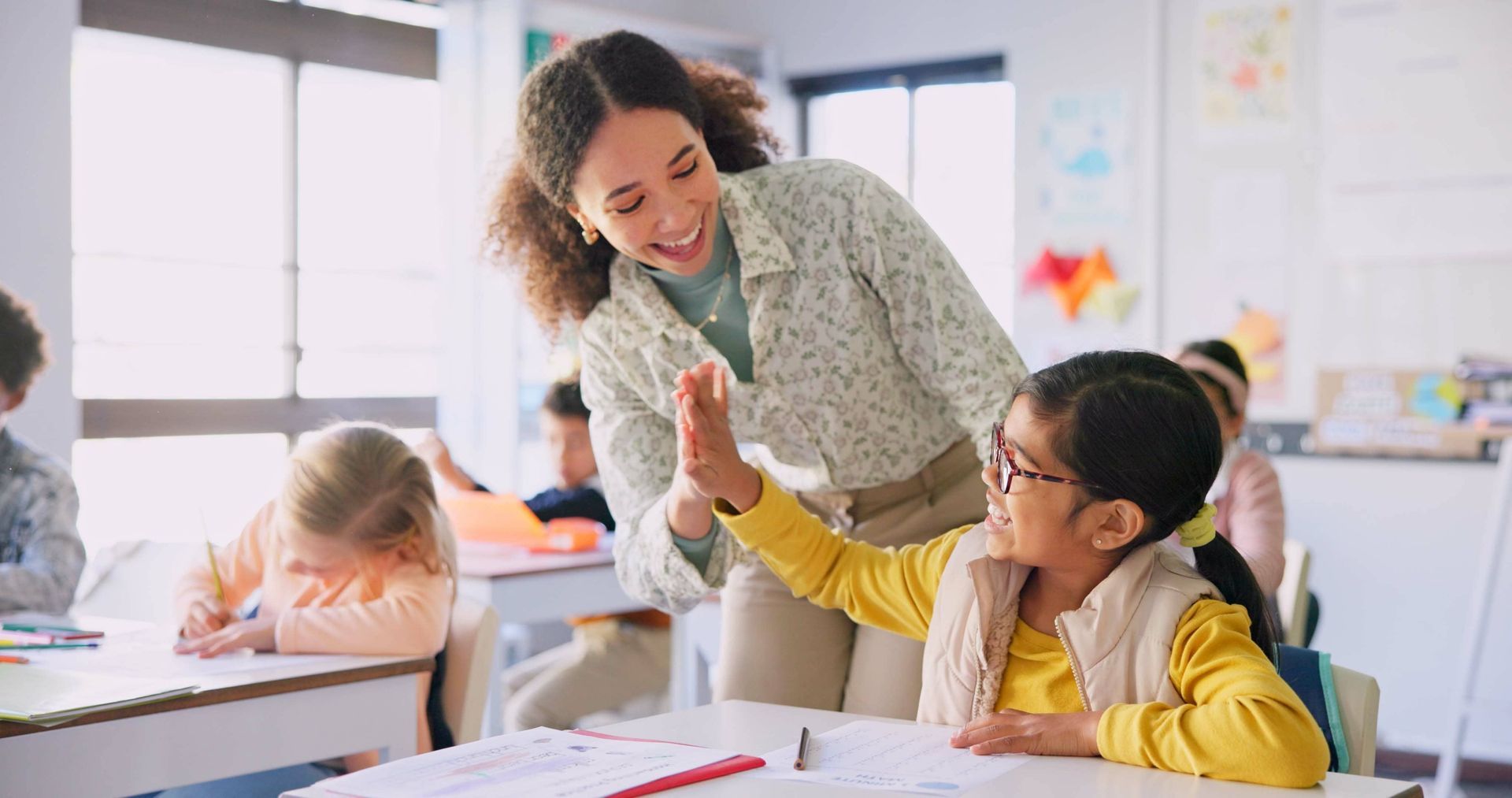 Teacher giving a high-five to a student in a classroom setting. Other children are seated at desks.