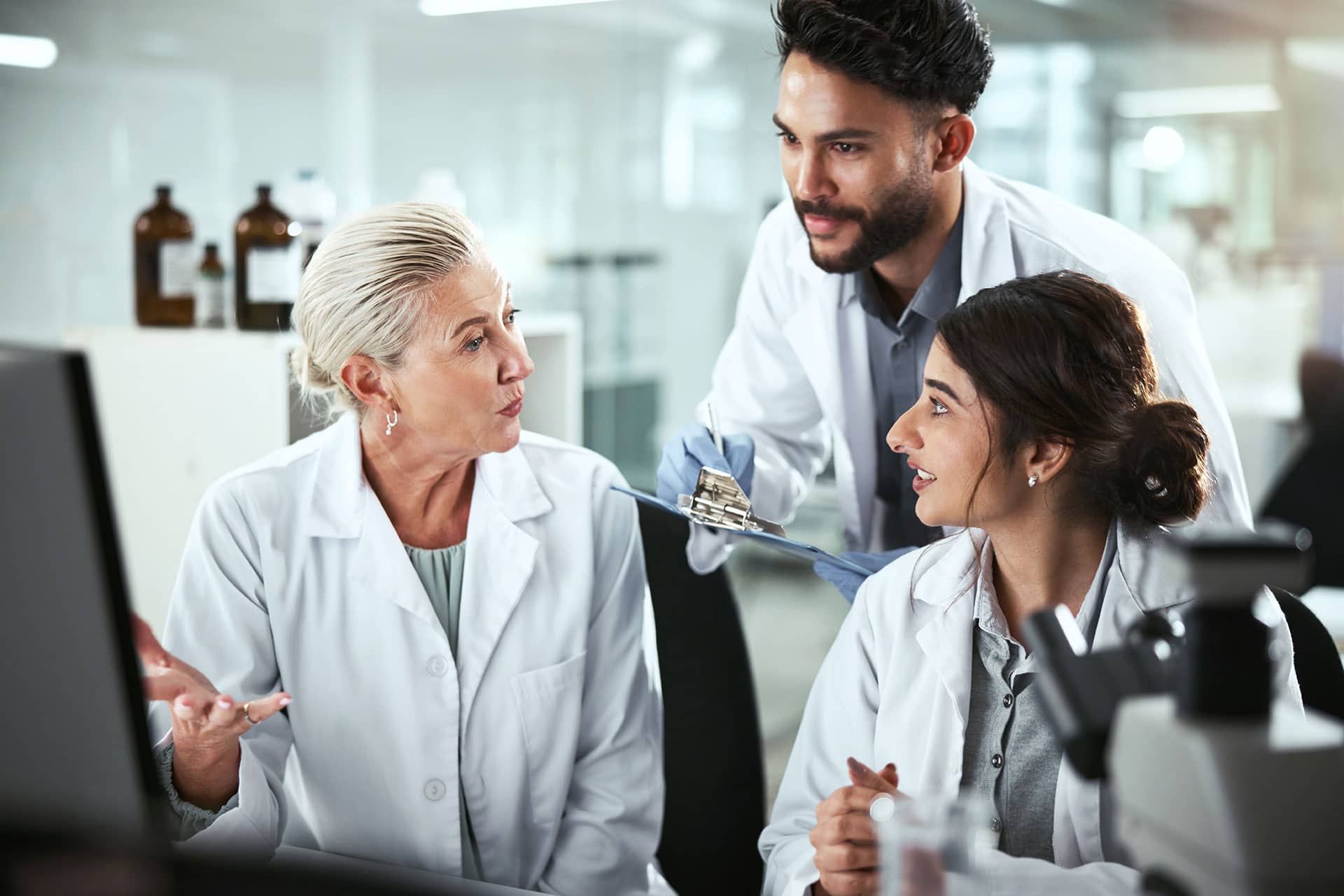 Three lab workers in white coats discussing results in a lab setting.