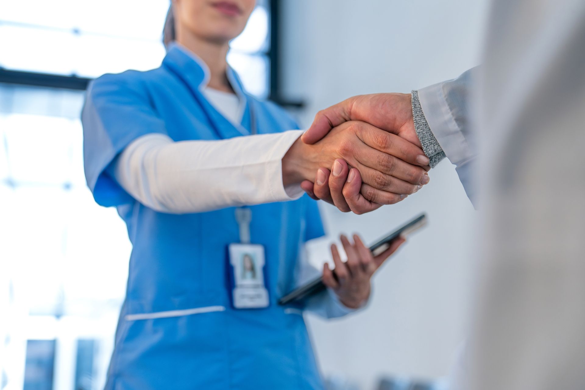 Operatori sanitari si stringono la mano in un ambiente medico. Uno indossa un camice blu e tiene in mano un tablet.