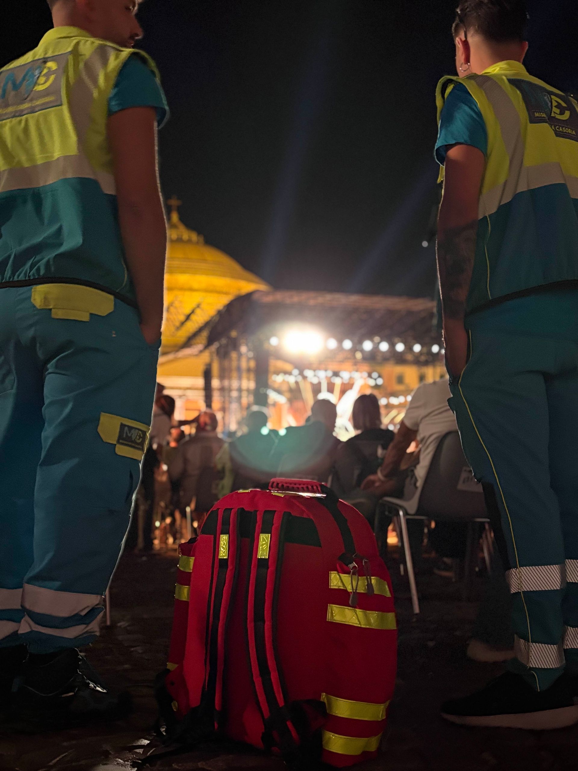 Due operatori sanitari sono in piedi accanto a una borsa rossa per le emergenze e osservano la folla di notte.