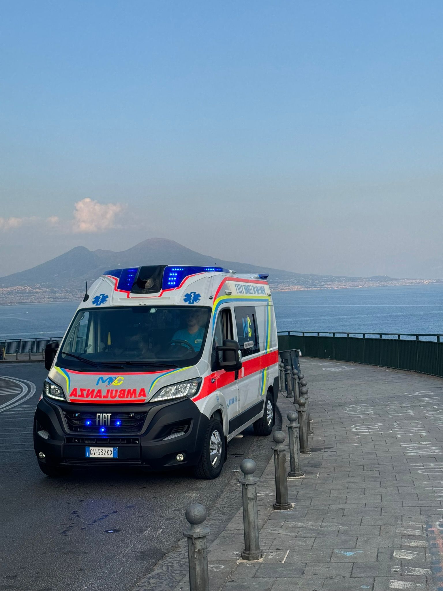 Ambulanza parcheggiata sul lungomare, luci blu accese, mare e montagna sullo sfondo.