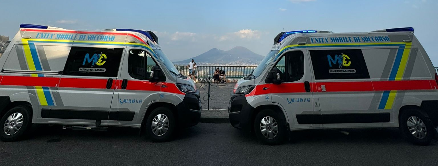 Due ambulanze parcheggiate una accanto all'altra, con strisce rosse e bianche, di fronte a una catena montuosa costiera.