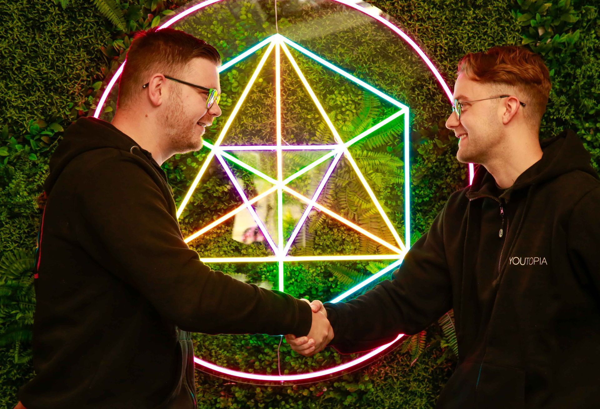 Two men shaking hands in front of a glowing geometric design on a green wall.