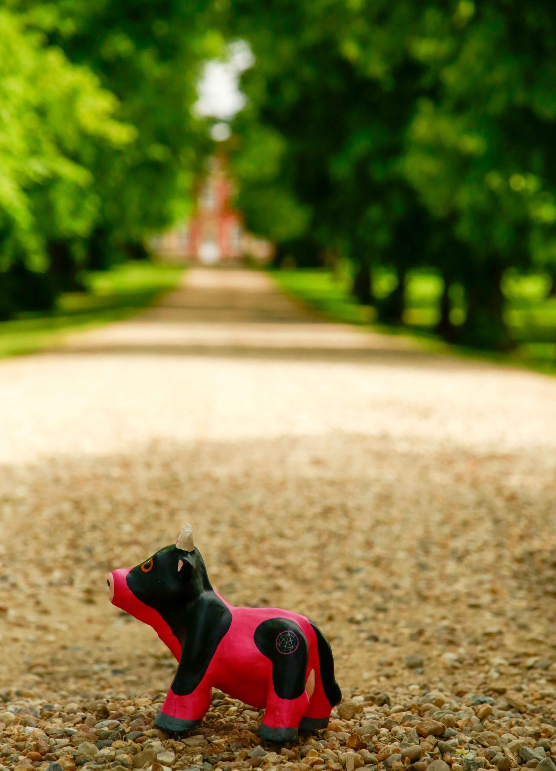 Toy cow on a gravel path, leading to a red building lined by trees.
