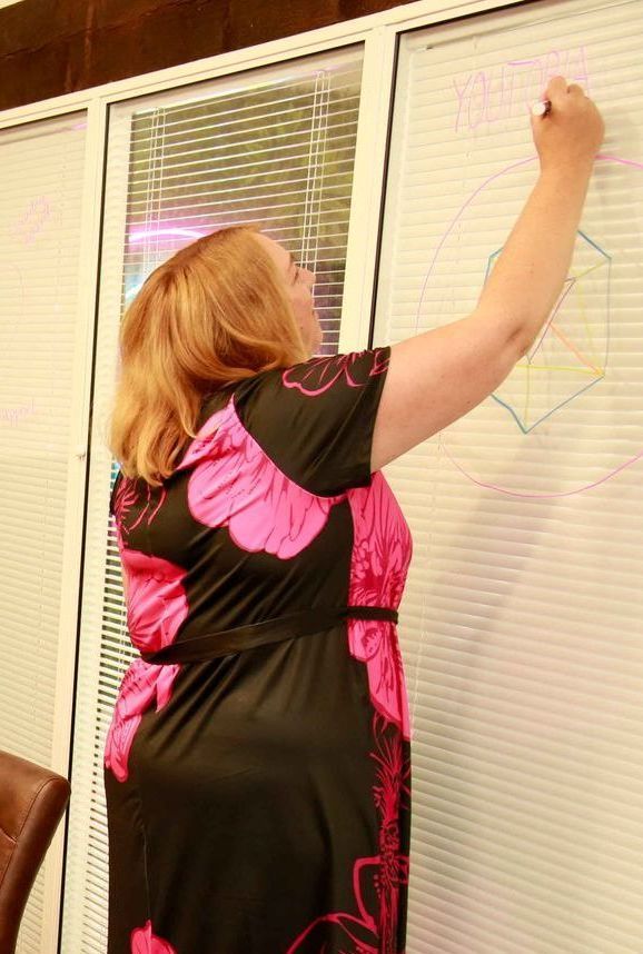 Woman in a black dress with pink floral design writing on a whiteboard, in front of a window with blinds.