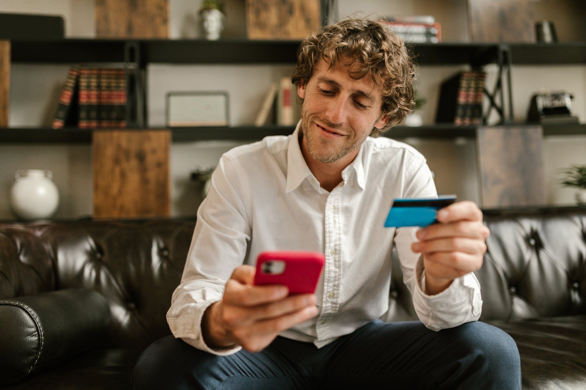 A man is sitting on a couch holding a credit card and a cell phone.
