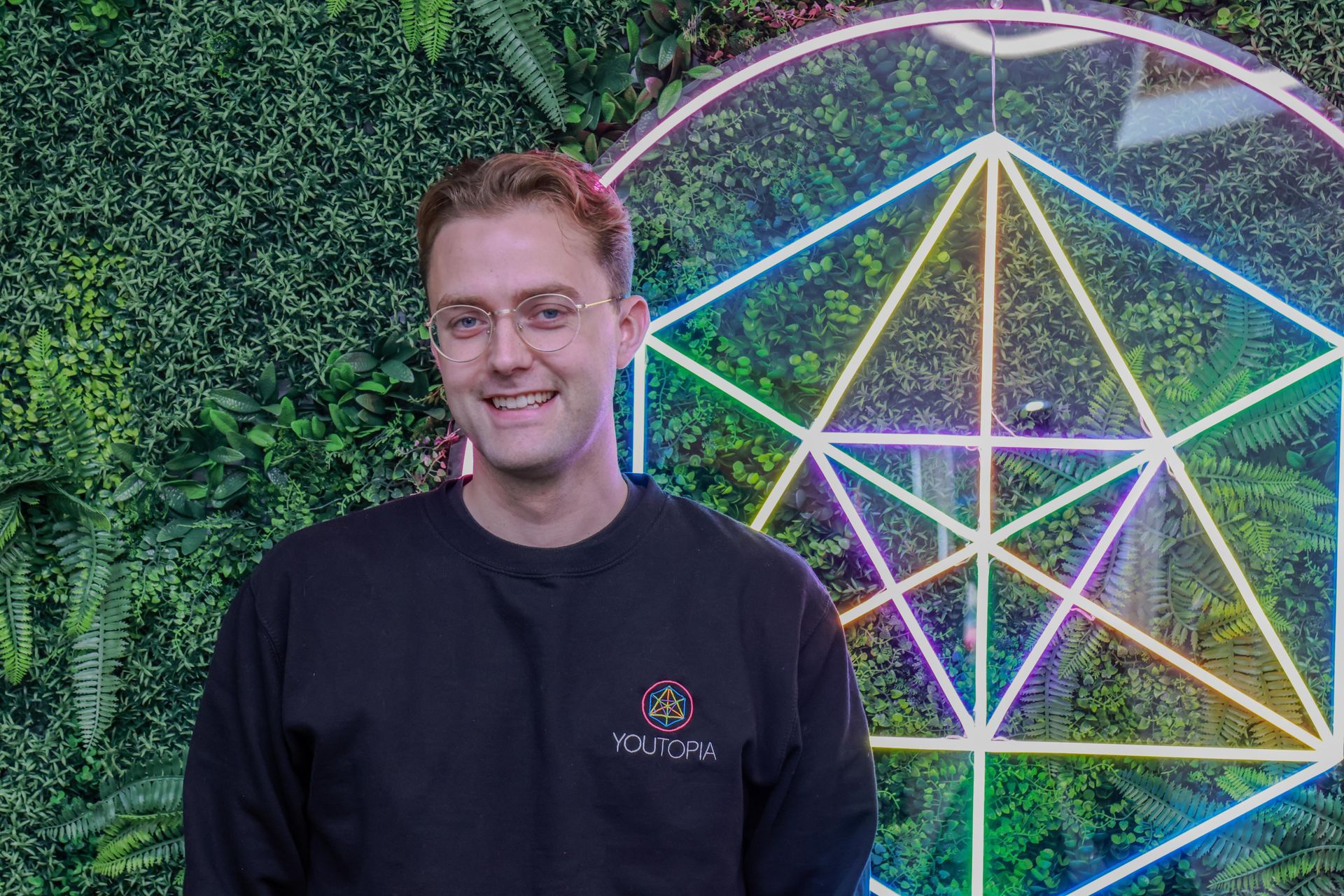 Smiling man in glasses beside a neon geometric light display on a green wall
