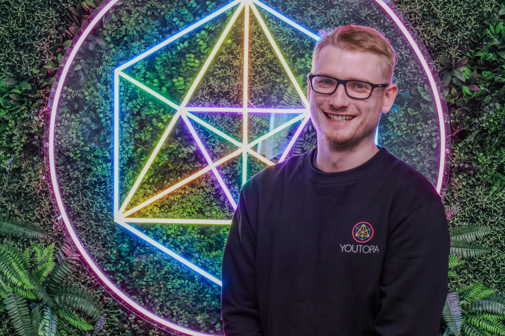 Smiling man in glasses before a neon geometric wall mural with a pentagram design
