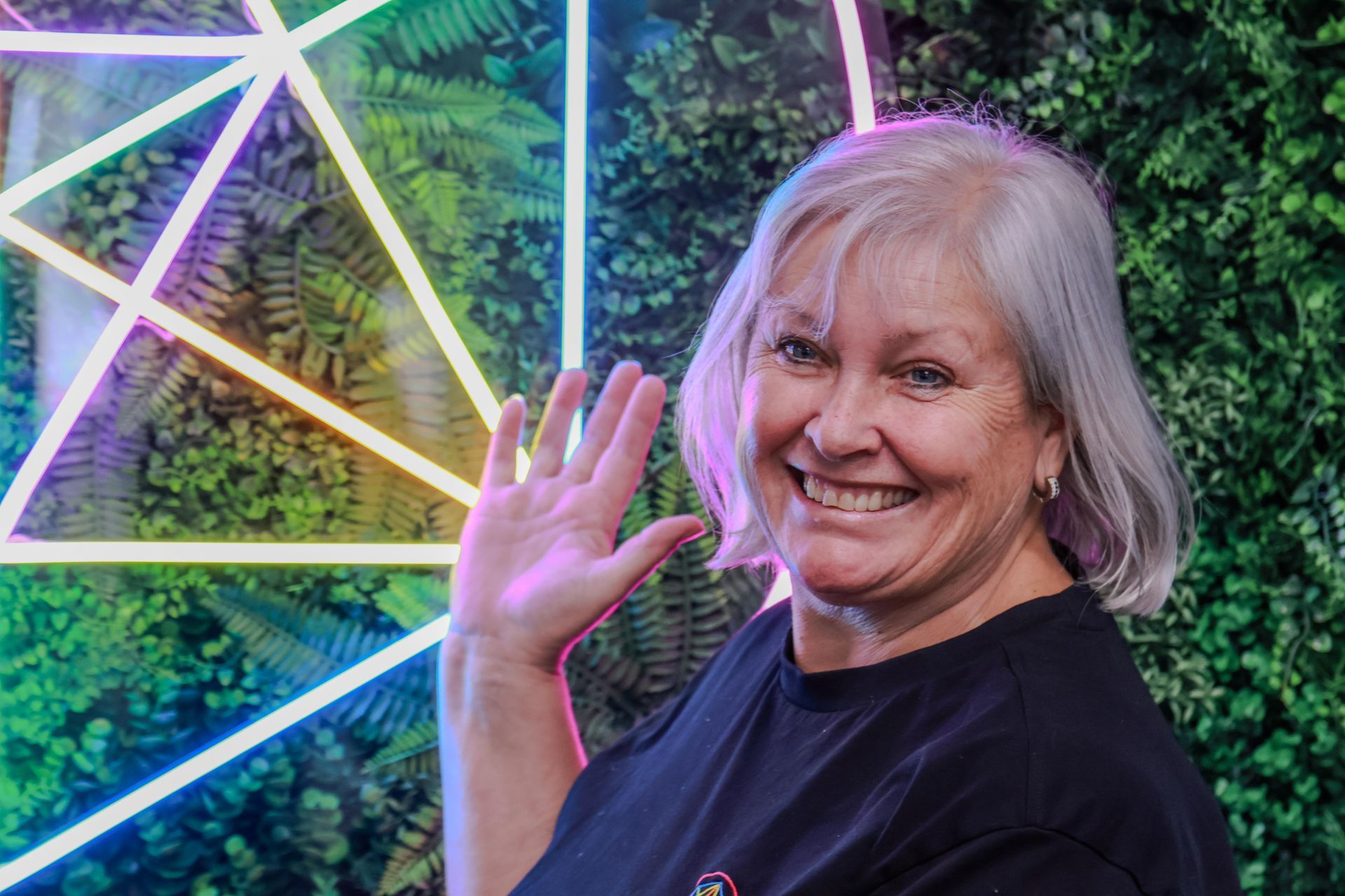 Smiling woman waves in front of colorful neon geometric lights outdoors