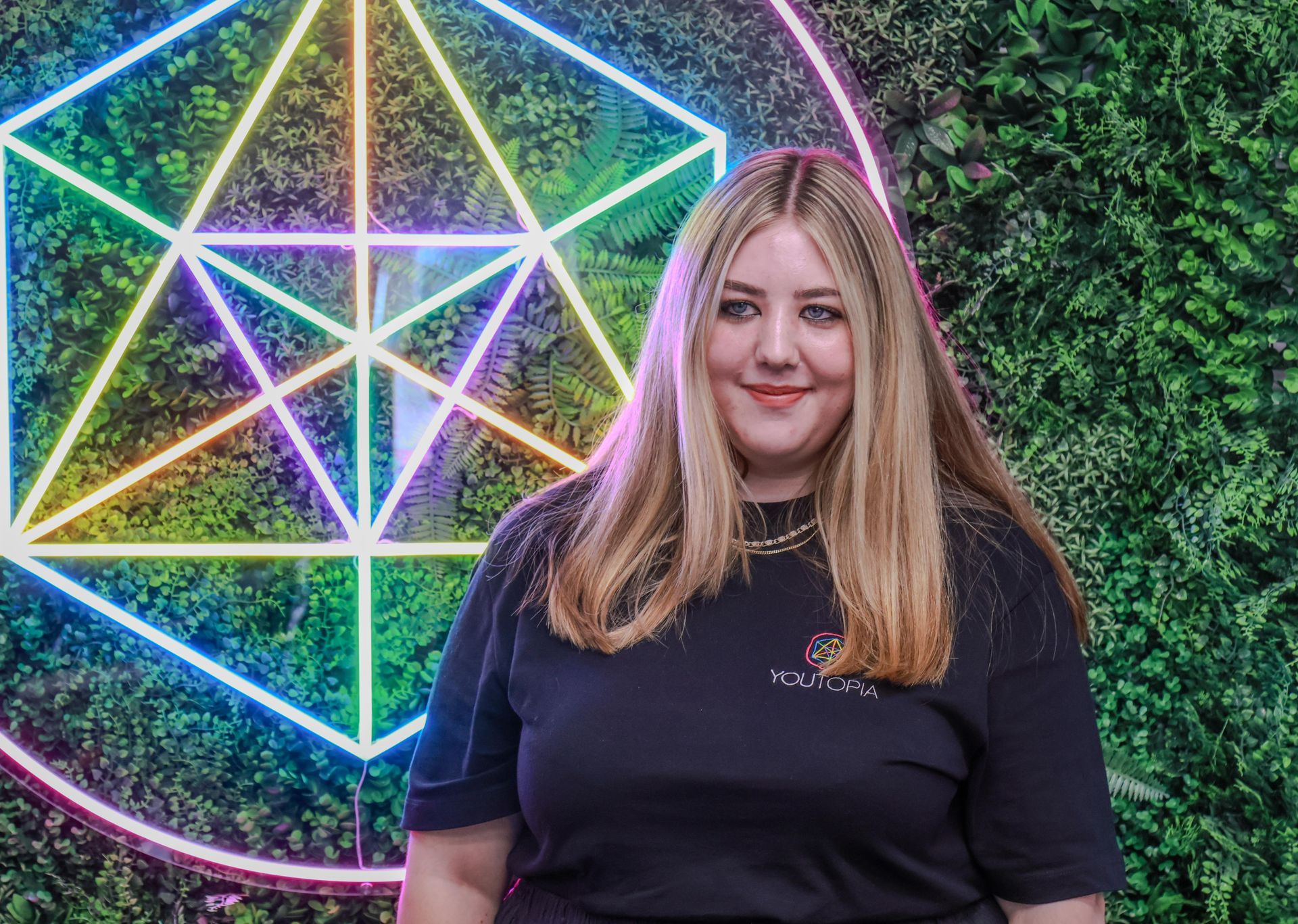 Woman standing in front of neon geometric lights and a green hedge wall, smiling at the camera.