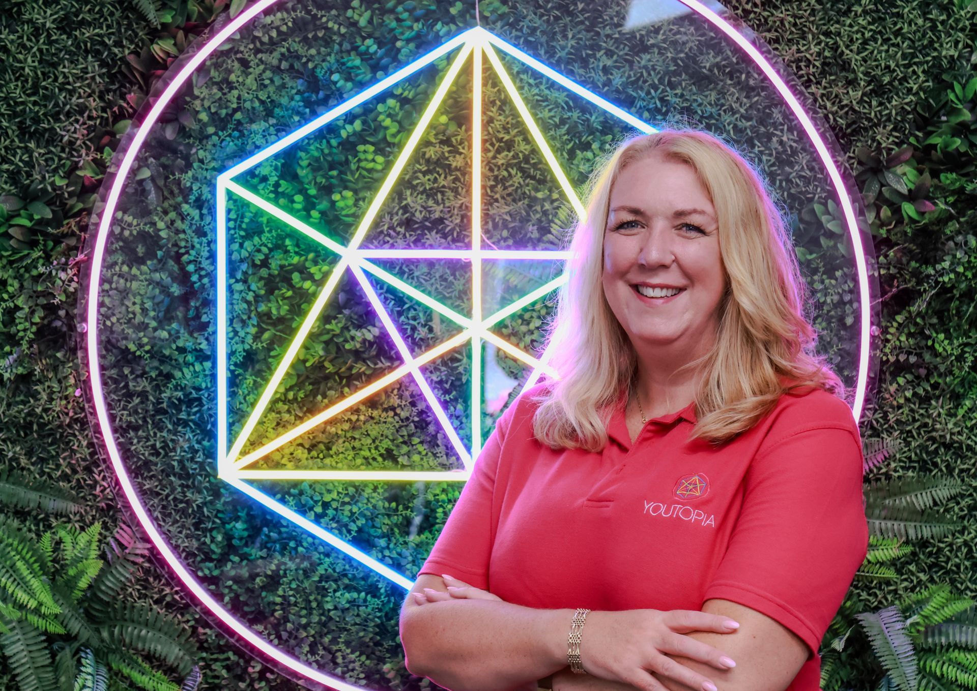Smiling woman in a coral shirt standing before a colorful geometric neon light installation outdoors