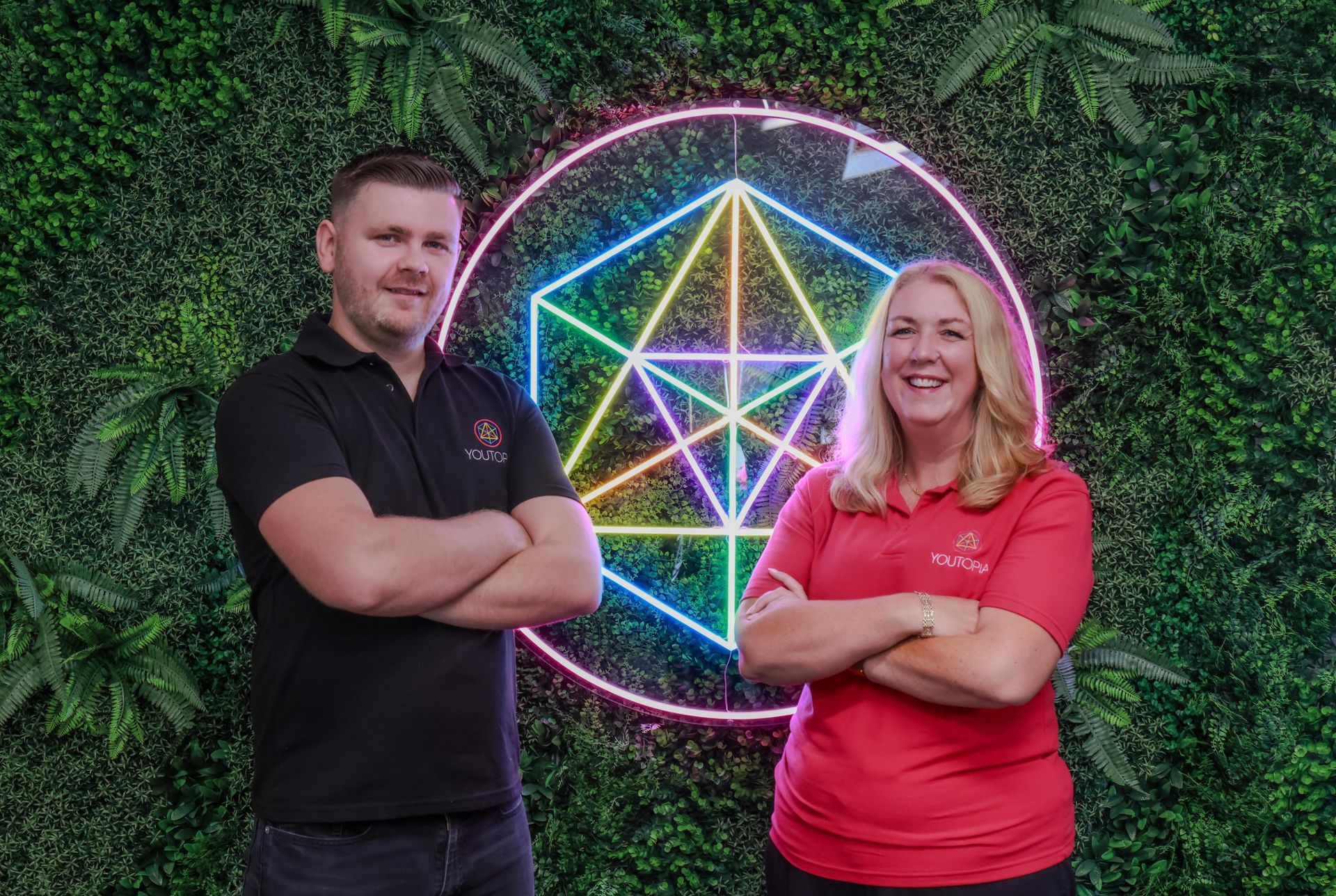 Two people with crossed arms in front of a neon geometric sign and a green wall.