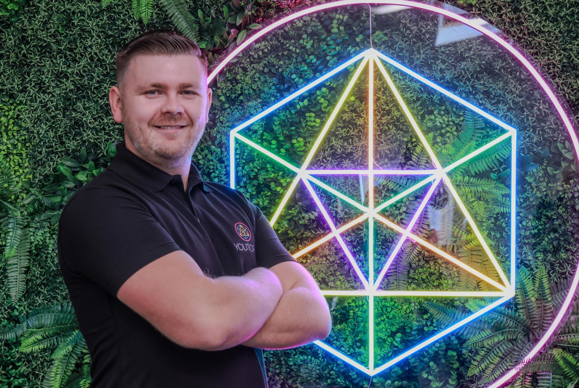 Man standing with arms crossed beside a neon geometric light display on a green wall