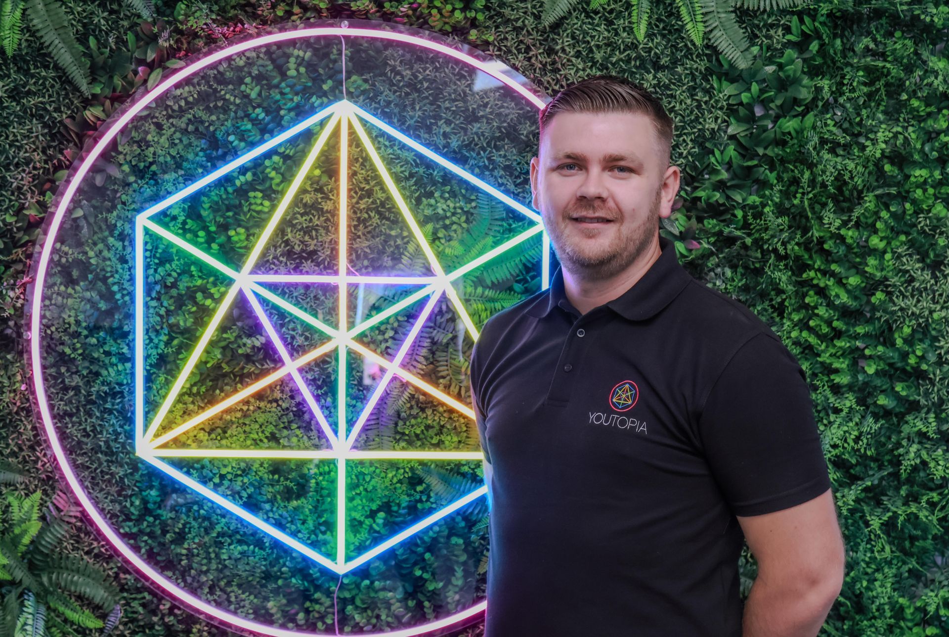 Man standing beside a neon geometric wall sign on a leafy green backdrop