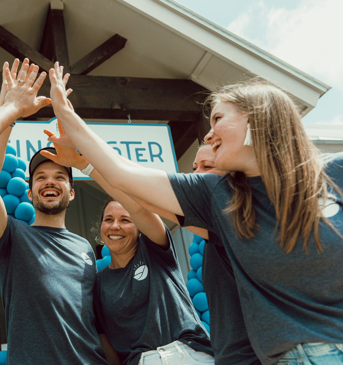 Three people in dark grey shirts smiling and reaching up to high-five under a blue balloon arch.