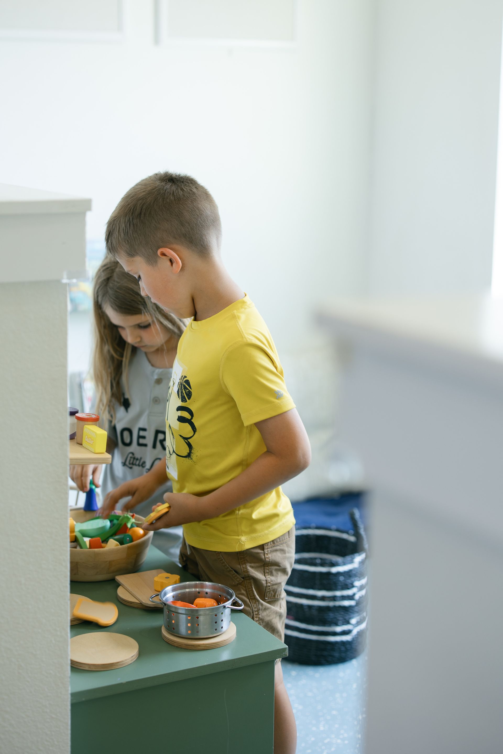 Two children playing with toy food at a child-sized counter, with a basket of vegetables.