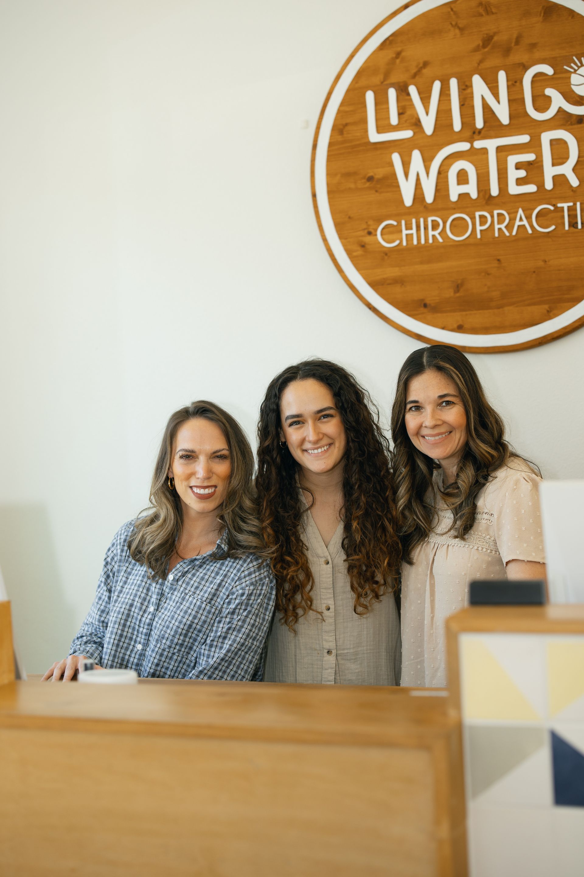 Three women smiling behind a wooden reception desk; Living Water Chiropractic sign on the wall.