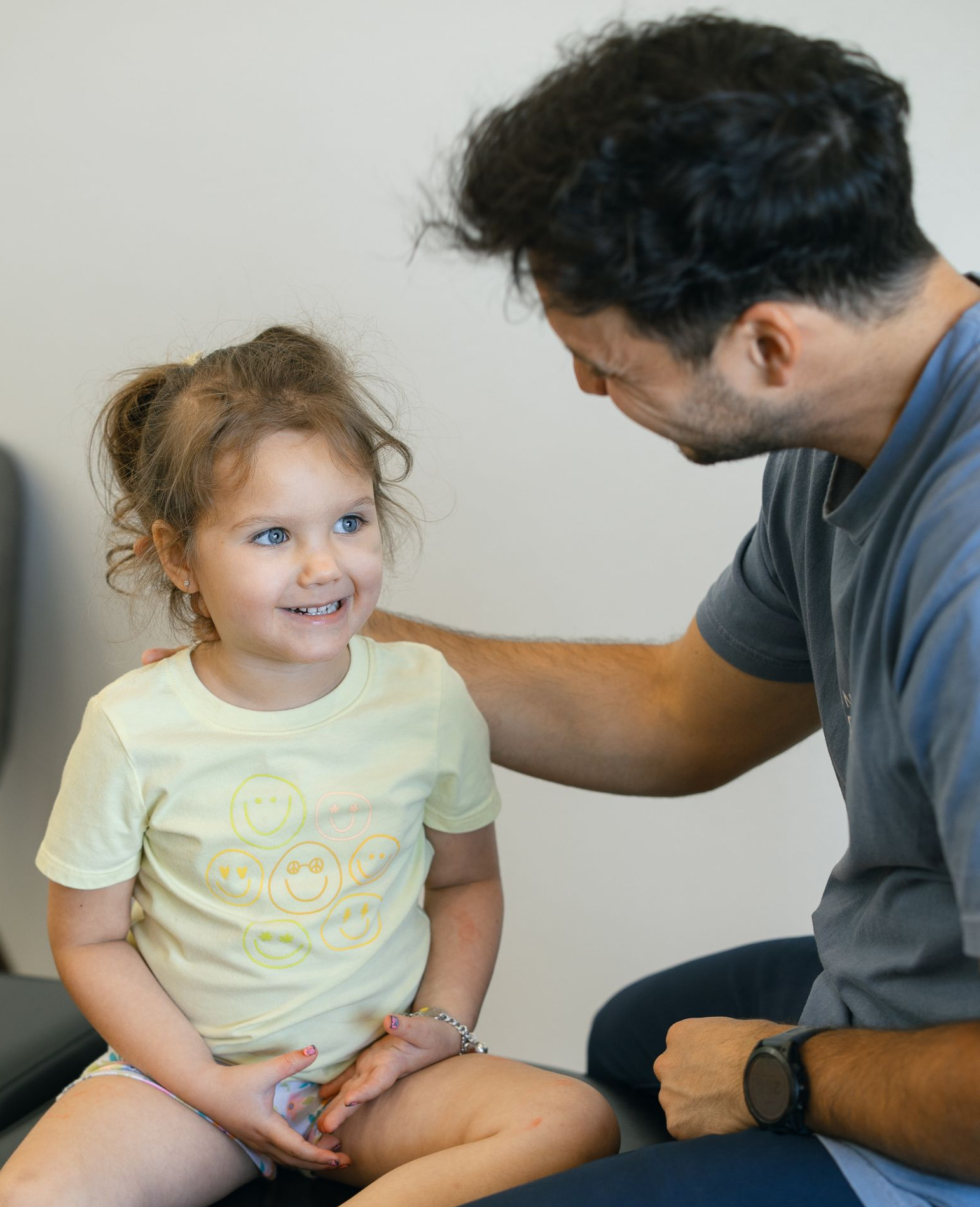 Adult talking with smiling child in a light shirt, seated together indoors on a couch.