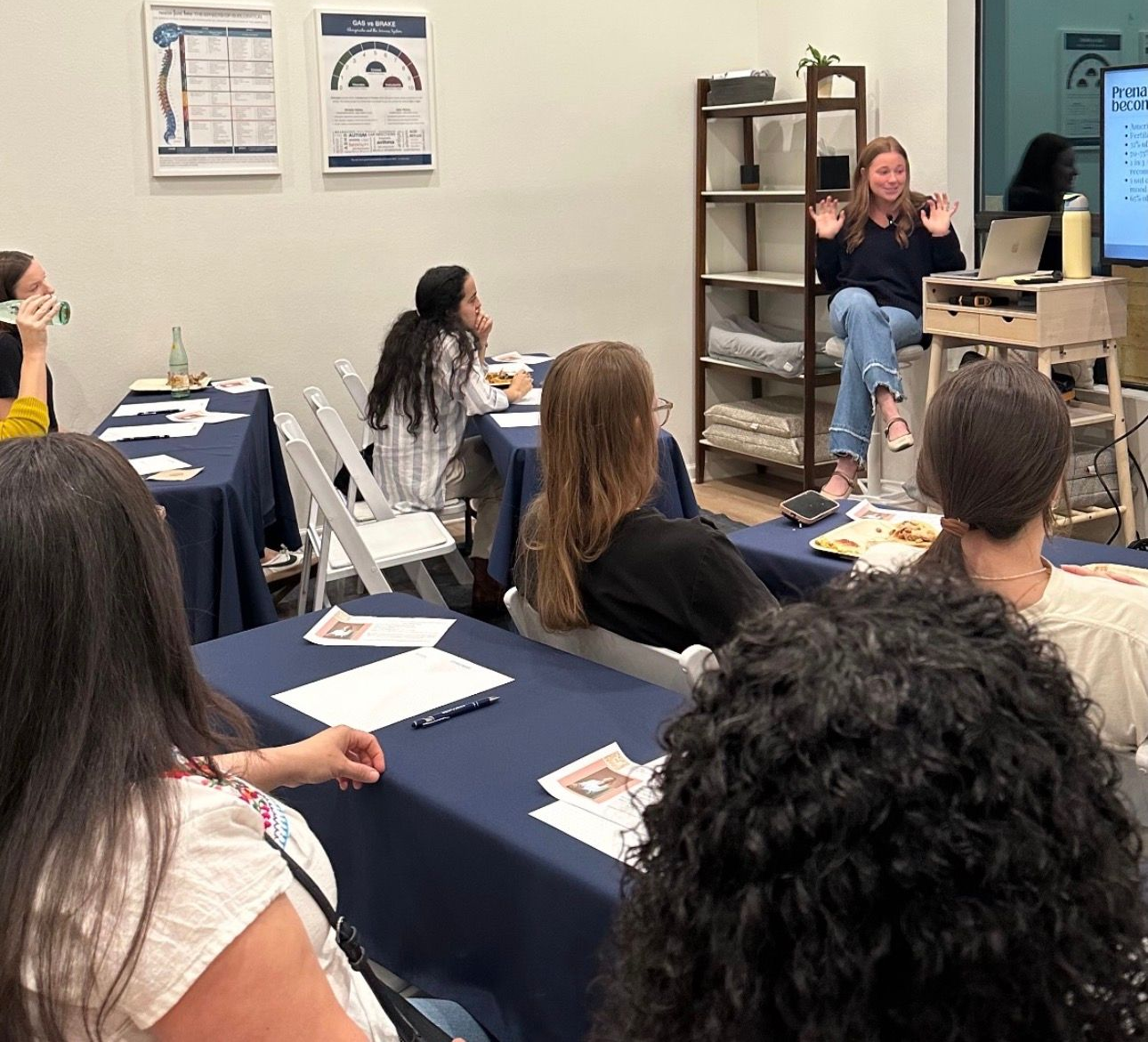 A speaker gestures during a presentation in a classroom setting, while an audience listens at tables with dark blue cloths.