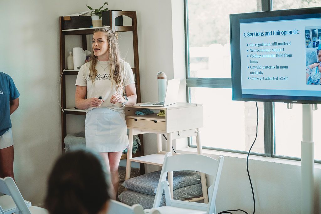 Woman presenting beside a screen in a classroom, with a seated audience and shelves in the background