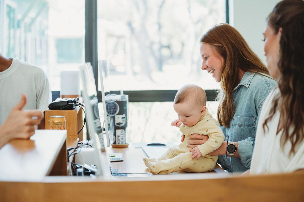 A woman holding a baby sits at a desk with an open laptop, smiling while another person is partially visible nearby.