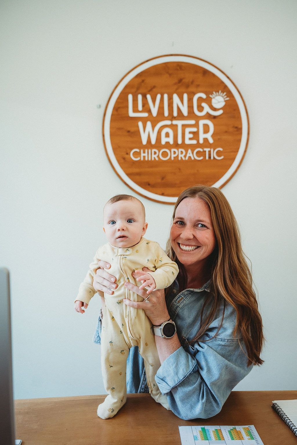 A person smiles while holding a baby in front of a circular wooden sign that reads