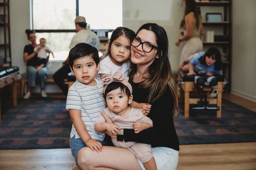 A smiling person holds a baby while two other children stand close by in a living room with others in the background.
