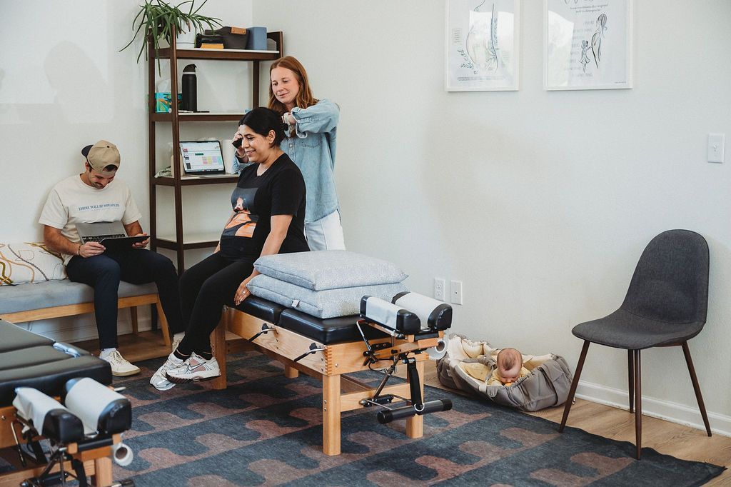A practitioner adjusts a seated client in a clinic while a person sits on a nearby bench and a dog rests on the floor.