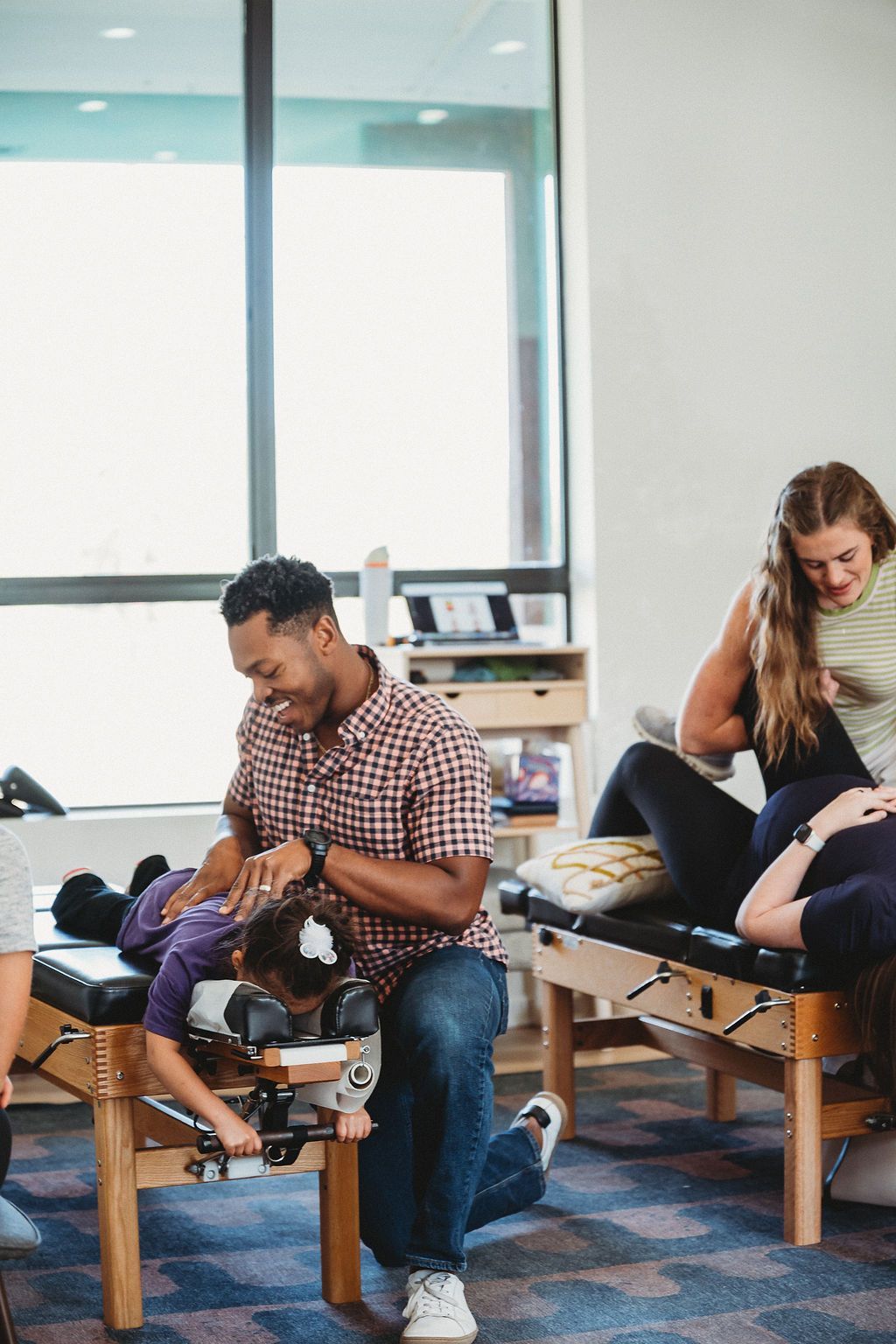 A chiropractor works on a patient lying on a treatment table in a clinic, with another person on a table in the background.