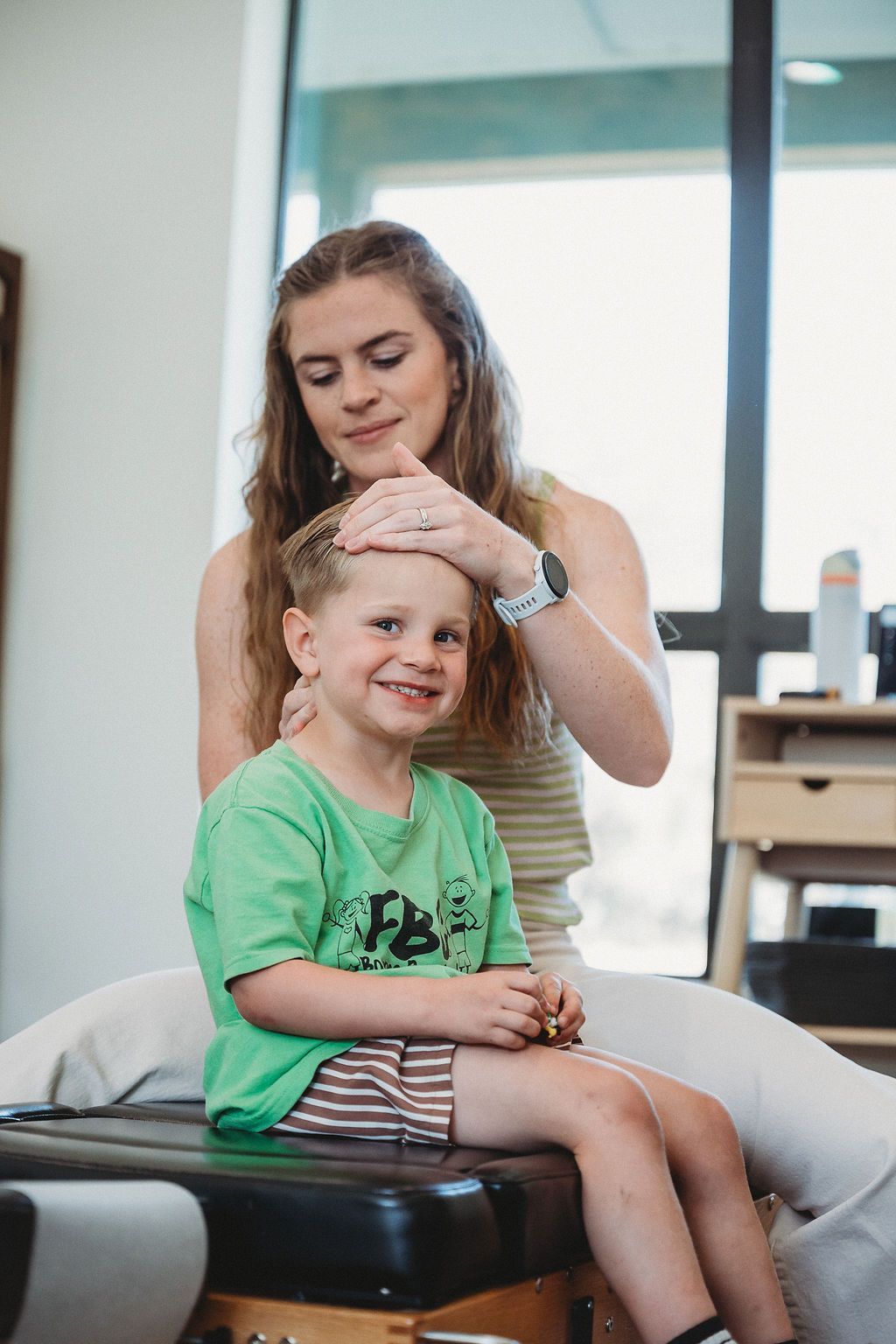 A practitioner gently places a hand on a smiling child’s head in a bright, clinical office setting.