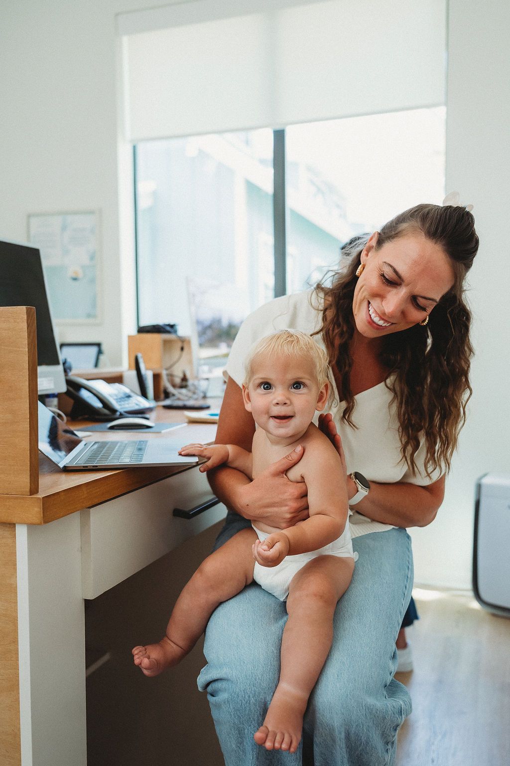 A parent smiles while working at a desk with a toddler sitting on their lap.