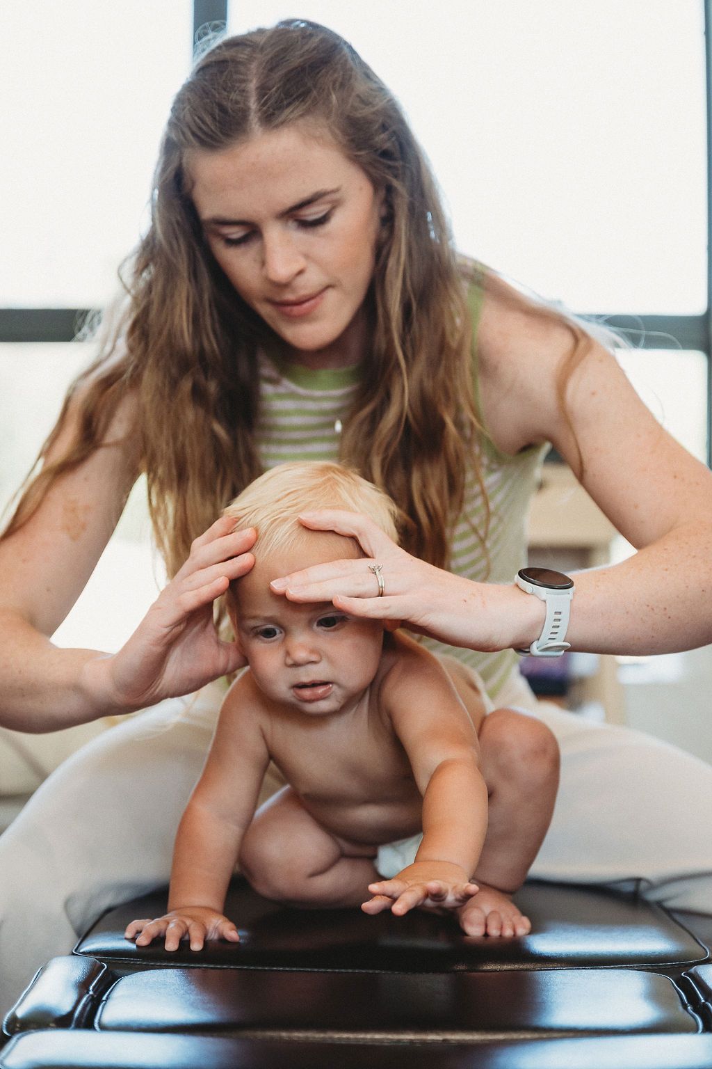 A person gently holds a toddler's head while the child crawls on a padded clinical table.