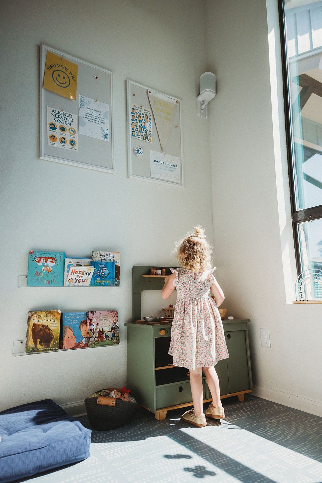 A child in a floral dress stands at a small green toy kitchen in a brightly lit room with wall-mounted book displays.