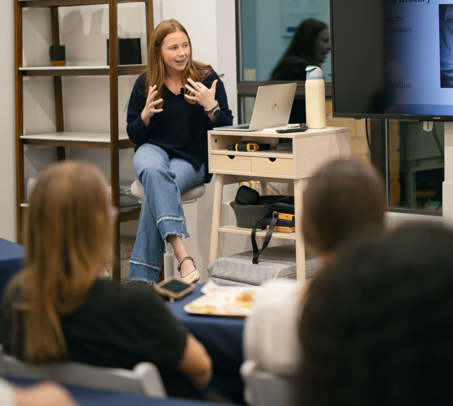 A speaker gestures while presenting to an audience seated in a bright, modern room with wooden shelves and a monitor.