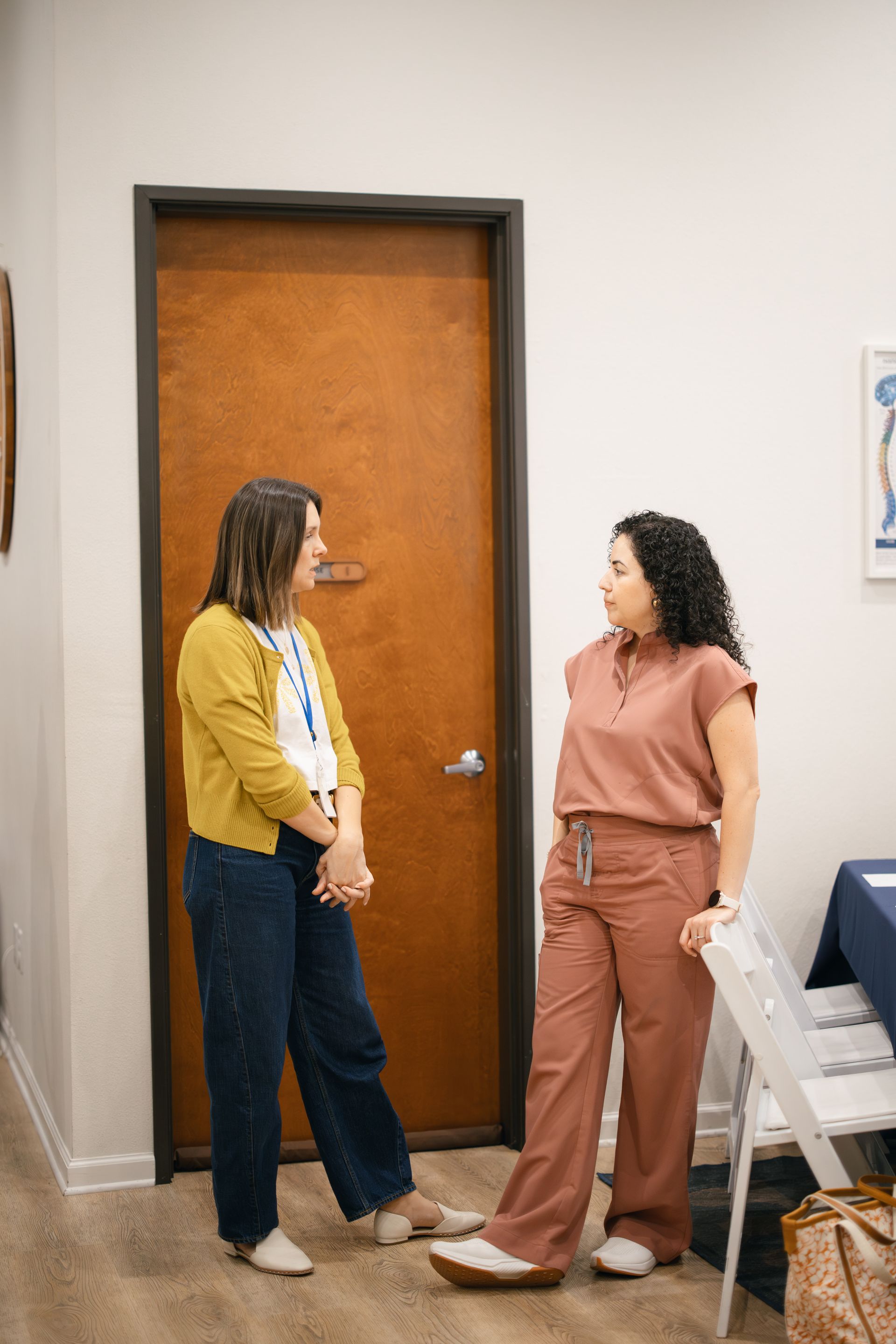 Two people stand in an office chatting in front of a wooden door. One wears a yellow cardigan, the other rusty-pink scrubs.