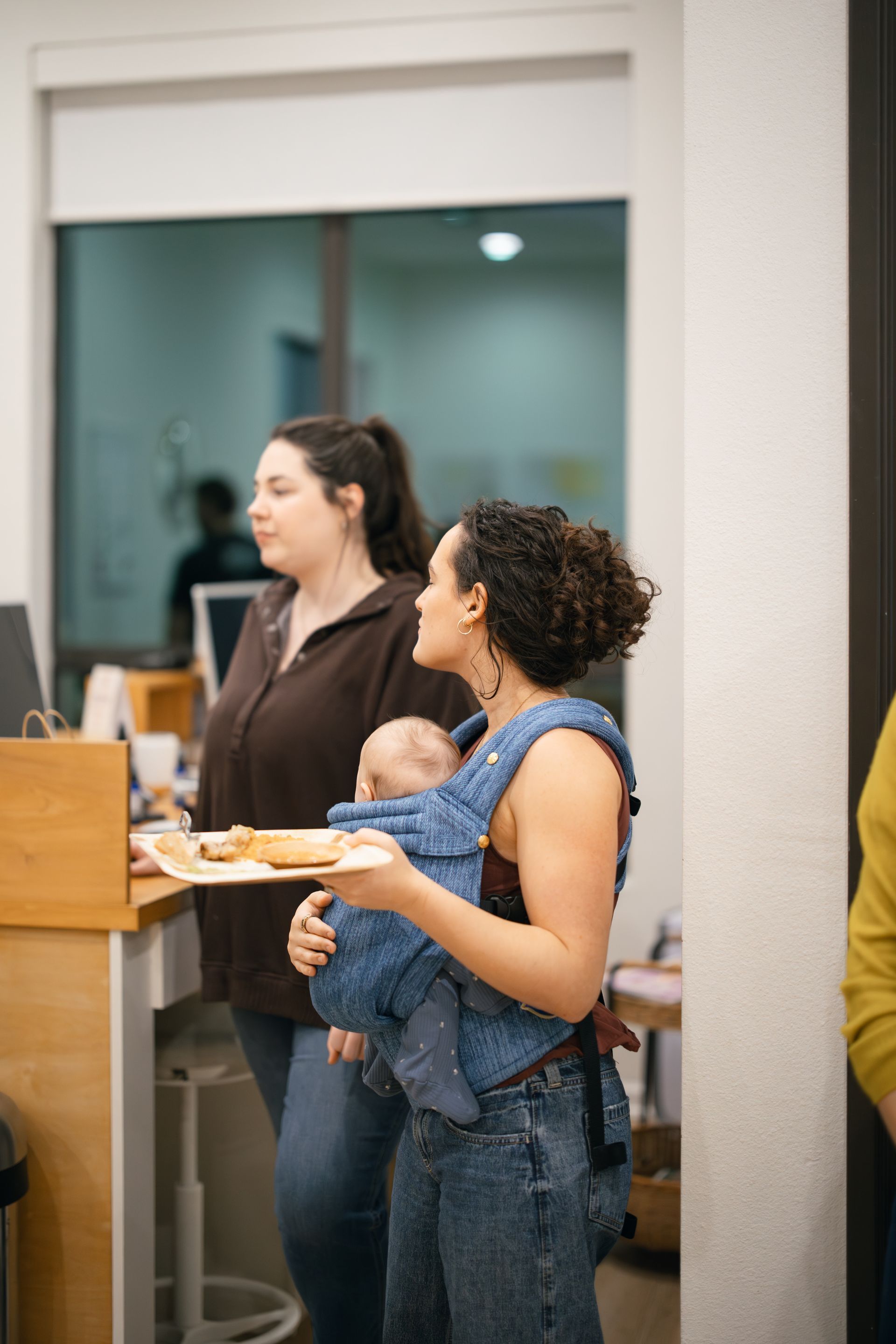 Two people stand in an indoor space, one wearing a baby in a blue carrier while holding a plate of food.