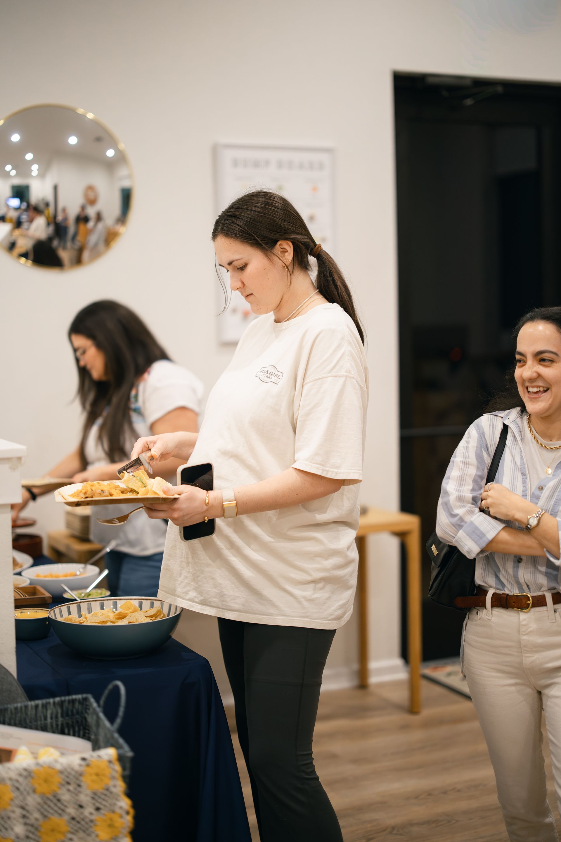 Three people gather at a food station with snacks, with one individual holding a plate, in a bright indoor setting.