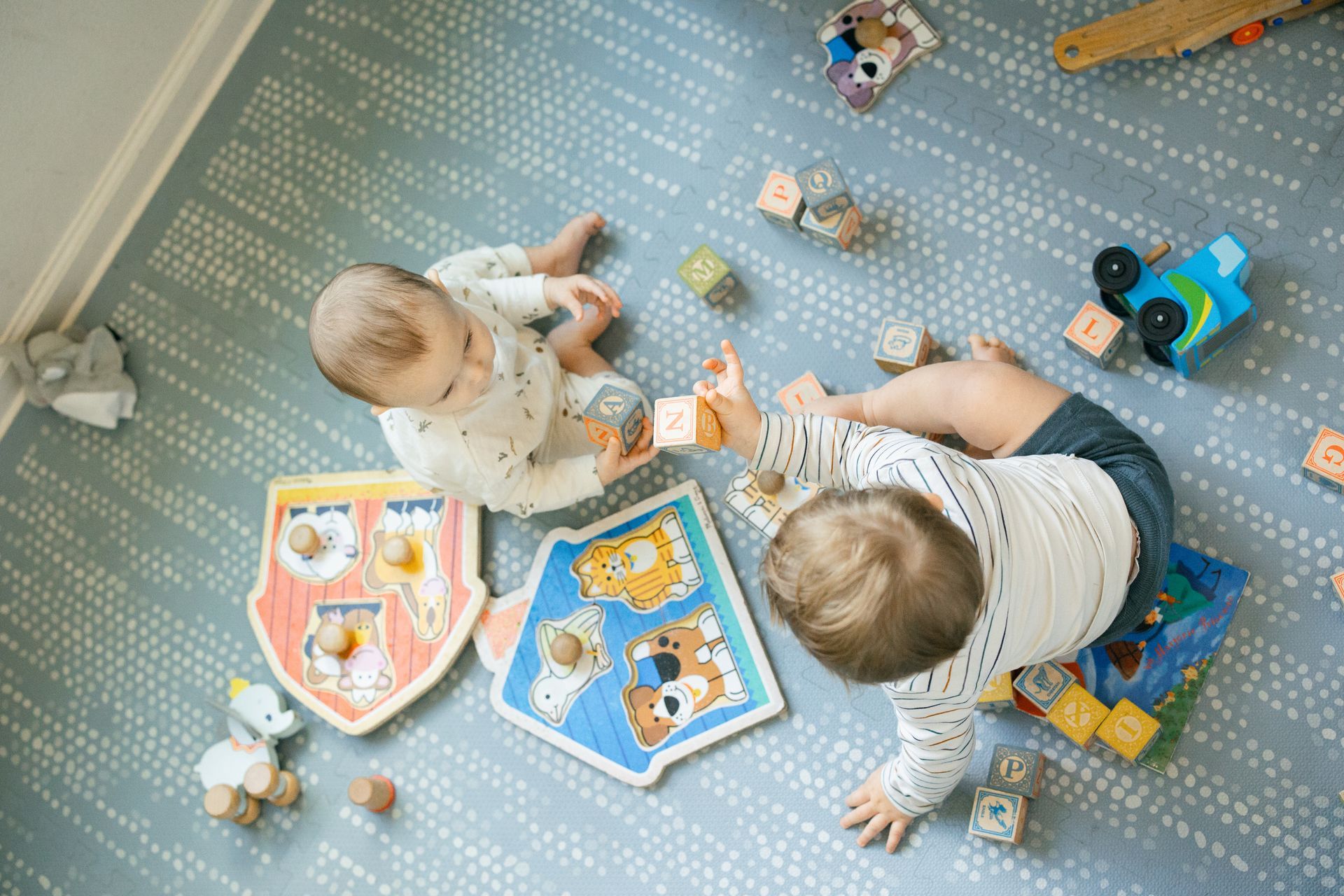 Two babies playing on a gray play mat with toys and colorful books around them